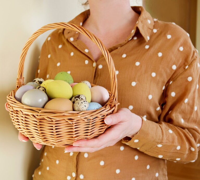 Nahaufnahme einer Frau in gepunkteter Bluse, die einen geflochtenen Korb mit dekorierten Ostereiern präsentiert. | © Adobe Stock/ agneskantaruk