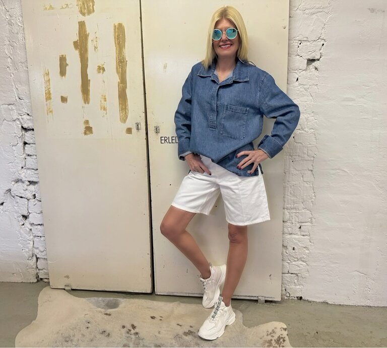 Mittelalte Frau mit glattem blondem Haar, Jeanstunika und runder Sonnenbrille posiert entspannt vor abgenutzter Tür. | © Renate Zott