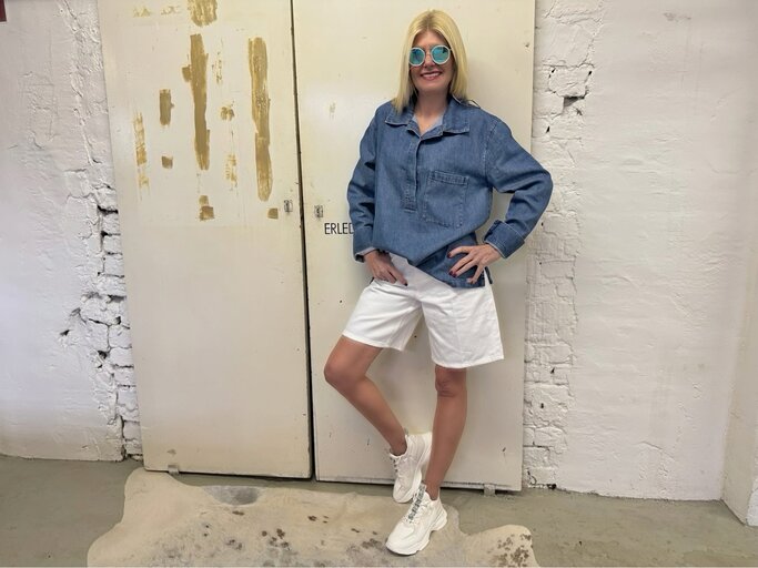 Mittelalte Frau mit glattem blondem Haar, Jeanstunika und runder Sonnenbrille posiert entspannt vor abgenutzter Tür. | © Renate Zott