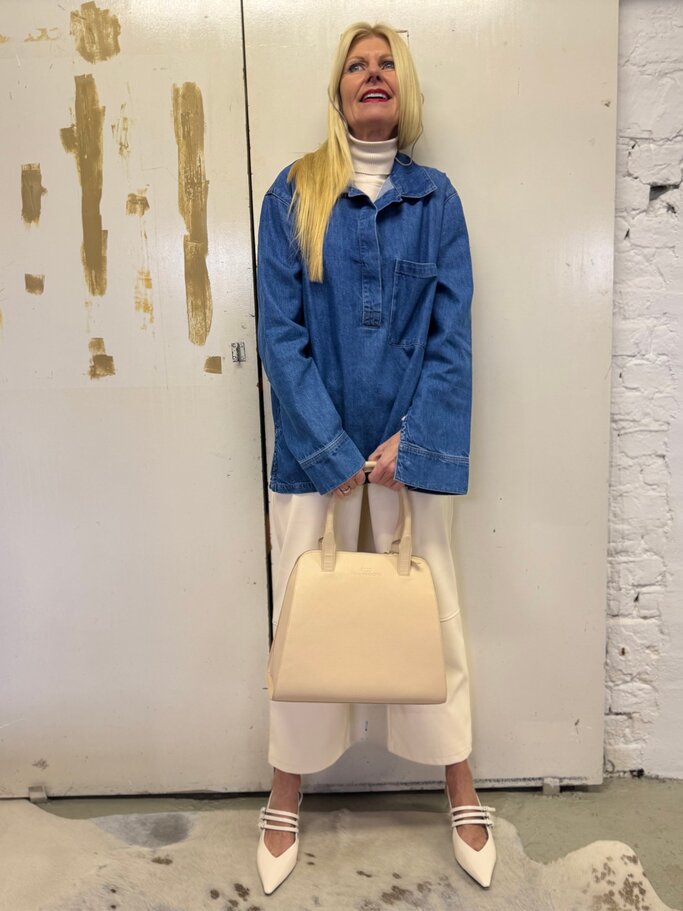 Modebewusste Frau in Oversized-Jeanstunika und weißem Outfit steht lächelnd mit beigefarbener Tasche vor abgenutzter Tür. | © Renate Zott