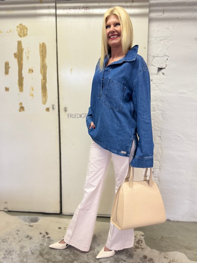 Modebewusste Frau in weiter Jeanstunika, weißen Hosen und beigen Accessoires steht entspannt vor heller Tür. | © Renate Zott