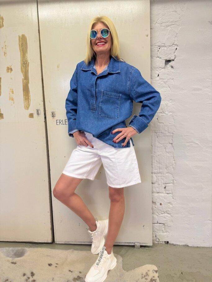 Stylishe Frau mittleren Alters trägt Jeanstunika, weiße Bermuda-Shorts und Sneaker, lehnt lächelnd vor heller Tür. | © Renate Zott