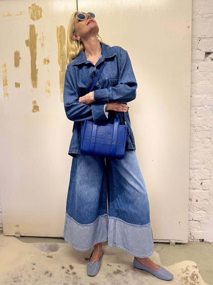 Blondhaarige Frau mit Jeanstunika, weiten Schlagjeans und blauer Tasche blickt lässig nach oben. | © Renate Zott