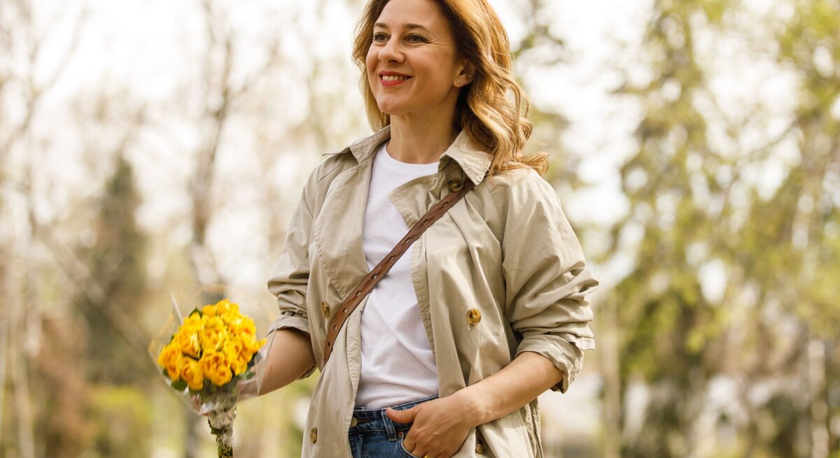 Zu sehen ist eine lächelnde Frau, die Rosen in der Hand hat und einen Trenchcoat trägt.