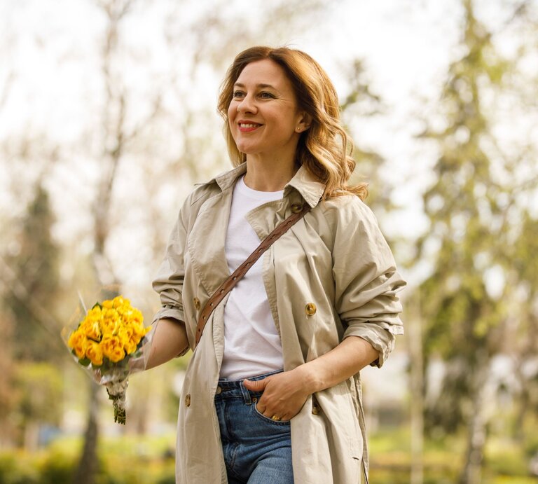 Zu sehen ist eine lächelnde Frau, die Rosen in der Hand hat und einen Trenchcoat trägt. | © GettyImages/fotostorm
