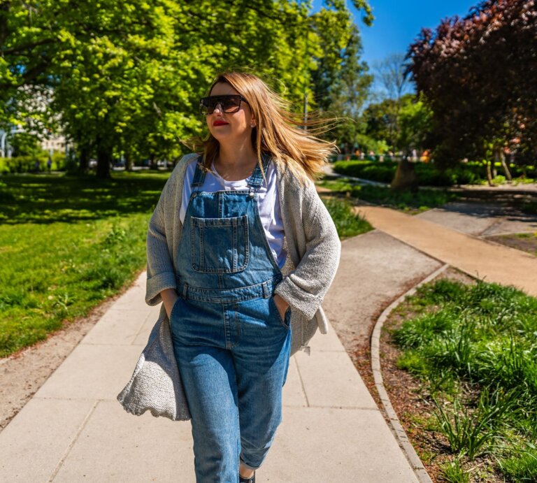 Frau in Latzhose und mit beiger Tasche genießt das Frühlingswetter in einem Park, umgeben von blühenden Bäumen und sattgrünem Gras. | © Adobe Stock/ Jacek Chabraszewski