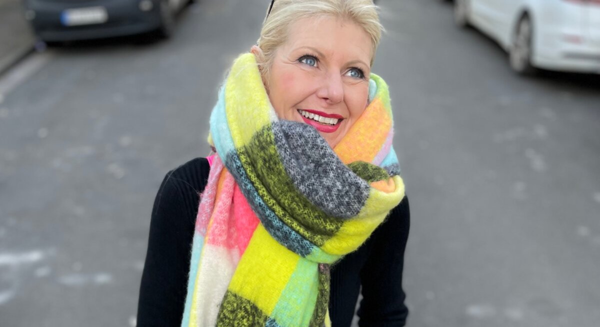 Renate Zott mit hochgestecktem Haar, buntem XXL-Schal und schwarzem Outfit steht auf einer Stadtstraße mit geparkten Autos.