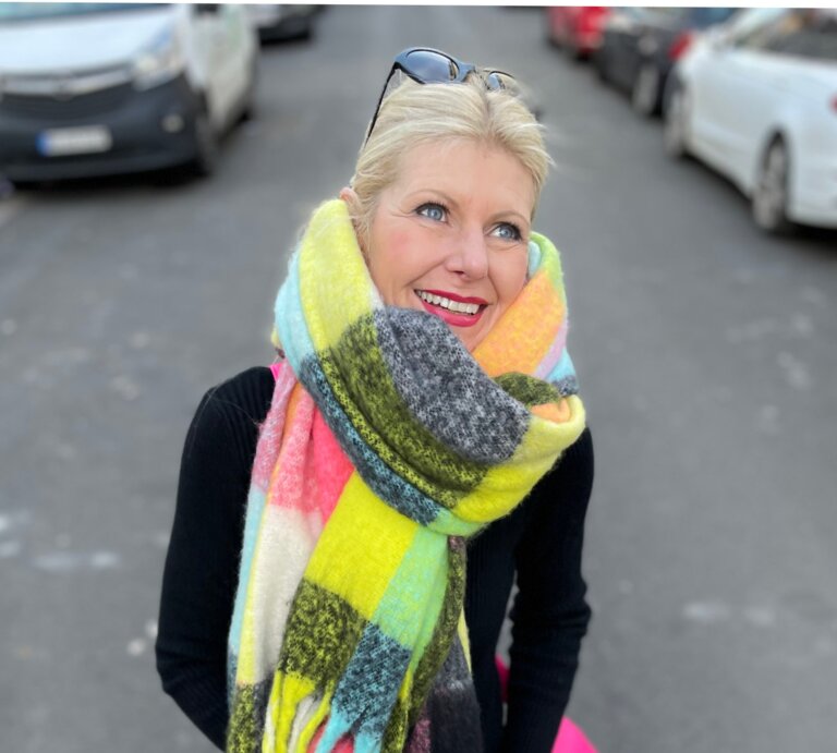 Renate Zott mit hochgestecktem Haar, buntem XXL-Schal und schwarzem Outfit steht auf einer Stadtstraße mit geparkten Autos. | © Renate Zott