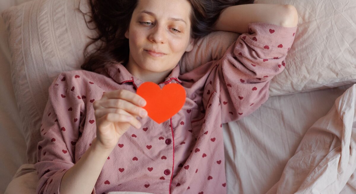 Eine Frau mit braunen Haaren liegt entspannt im Bett, trägt einen rosa Pyjama mit Herzmuster und hält ein rotes Papierherz in der Hand.