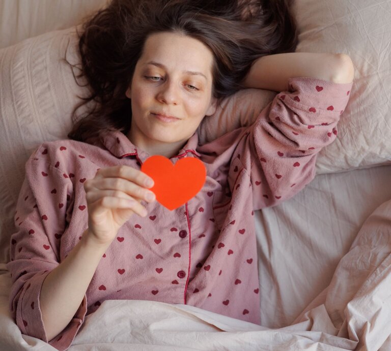 Eine Frau mit braunen Haaren liegt entspannt im Bett, trägt einen rosa Pyjama mit Herzmuster und hält ein rotes Papierherz in der Hand. | © Adobe Stock/ MyJuly