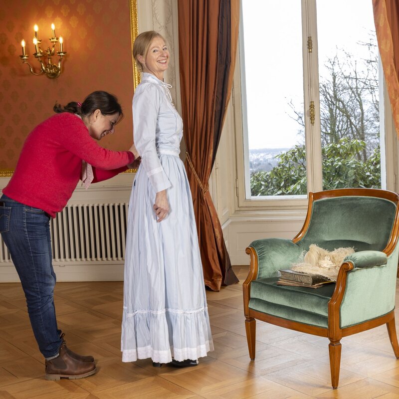 Eine Frau in historischer Kleidung wird von einer Assistentin angekleidet, neben ihnen ein grüner Sessel mit Hut und Büchern. | © Bernd Hanselmann