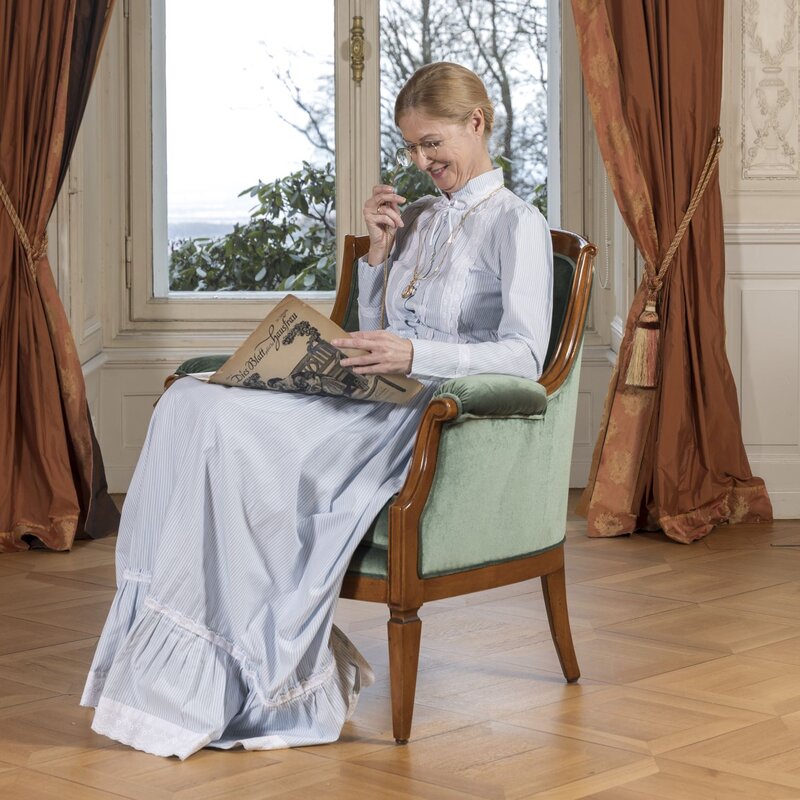 Eine Frau in hellblauem historischem Kleid sitzt lächelnd in einem grünen Sessel und liest eine alte Zeitung. | © Bernd Hanselmann