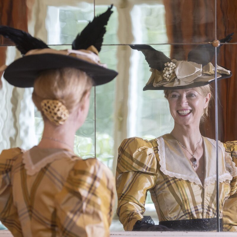 Eine Dame im historischen Kleid betrachtet sich mit einem strahlenden Lächeln im Spiegel, ihr kunstvoller Hut mit Federn im Fokus. | © Bernd Hanselmann