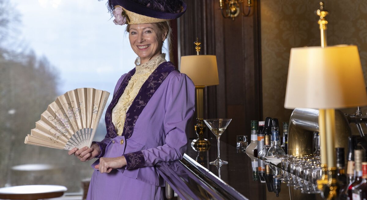 Eine elegant gekleidete Frau in violettem Kleid mit Spitzenkragen lehnt lächelnd an einer Bar und hält einen Fächer.