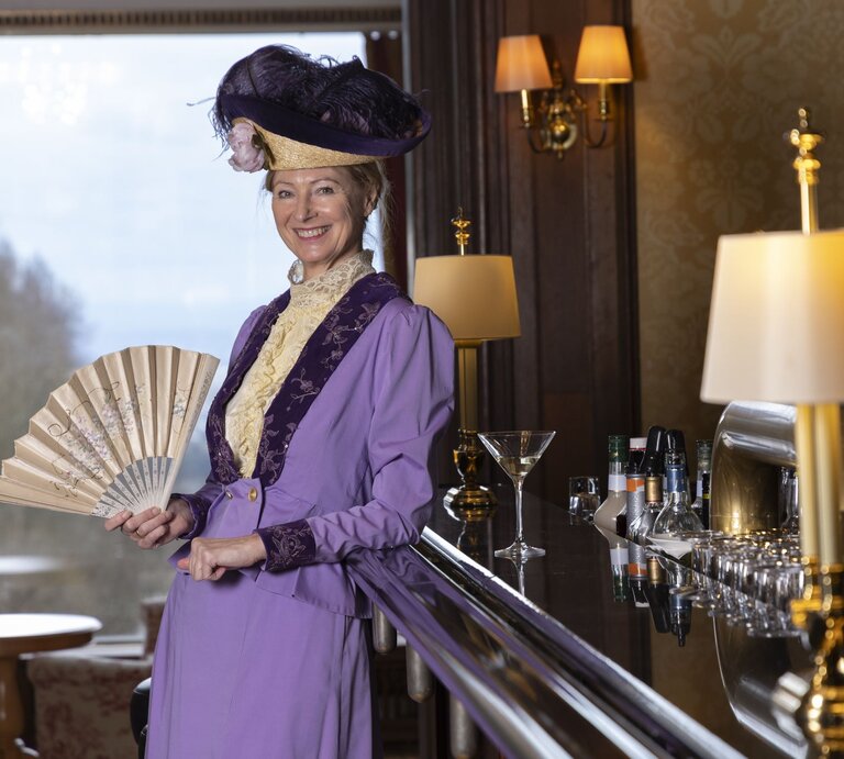 Eine elegant gekleidete Frau in violettem Kleid mit Spitzenkragen lehnt lächelnd an einer Bar und hält einen Fächer. | © Bernd Hanselmann