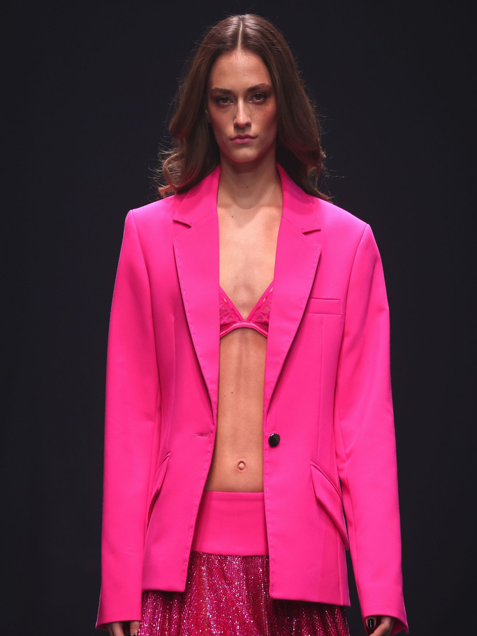 Model mit welligem Haar trägt einen oversized pinken Blazer über einem glitzernden Minirock und Spitzen-Bralette auf dem Laufsteg. | © Sebastian Reuter