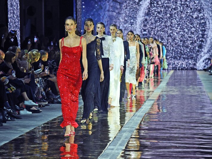 Eine Gruppe Models läuft in eleganten Abendkleidern über einen glänzenden Laufsteg, vorne eine Frau in funkelndem roten Paillettenkleid. | © Tristar Media/WireImage