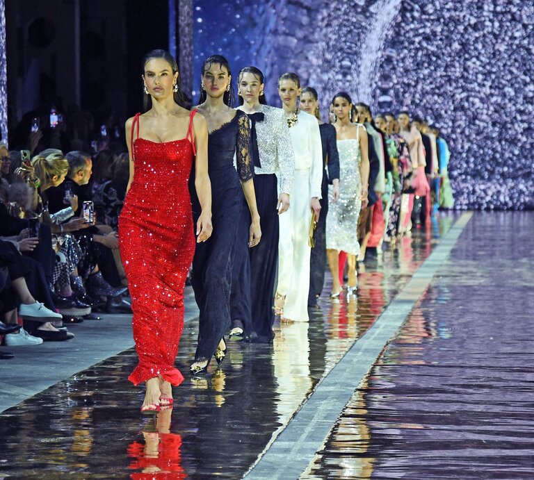 Eine Gruppe Models läuft in eleganten Abendkleidern über einen glänzenden Laufsteg, vorne eine Frau in funkelndem roten Paillettenkleid. | © Tristar Media/WireImage