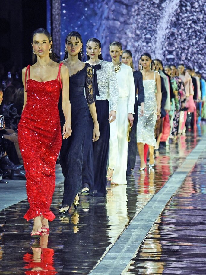 Eine Gruppe Models läuft in eleganten Abendkleidern über einen glänzenden Laufsteg, vorne eine Frau in funkelndem roten Paillettenkleid. | © Tristar Media/WireImage