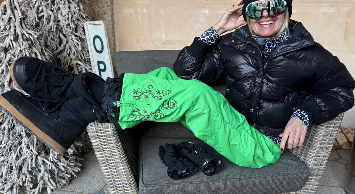 Renate Zott in schwarzer Daunenjacke, grüner Schneehose und dicker Mütze sitzt leger auf einem Korbstuhl, ein Handschuhpaar liegt daneben.