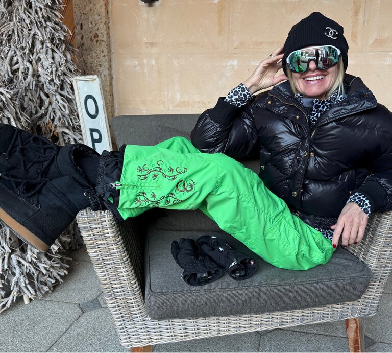 Renate Zott in schwarzer Daunenjacke, grüner Schneehose und dicker Mütze sitzt leger auf einem Korbstuhl, ein Handschuhpaar liegt daneben. | © Renate Zott