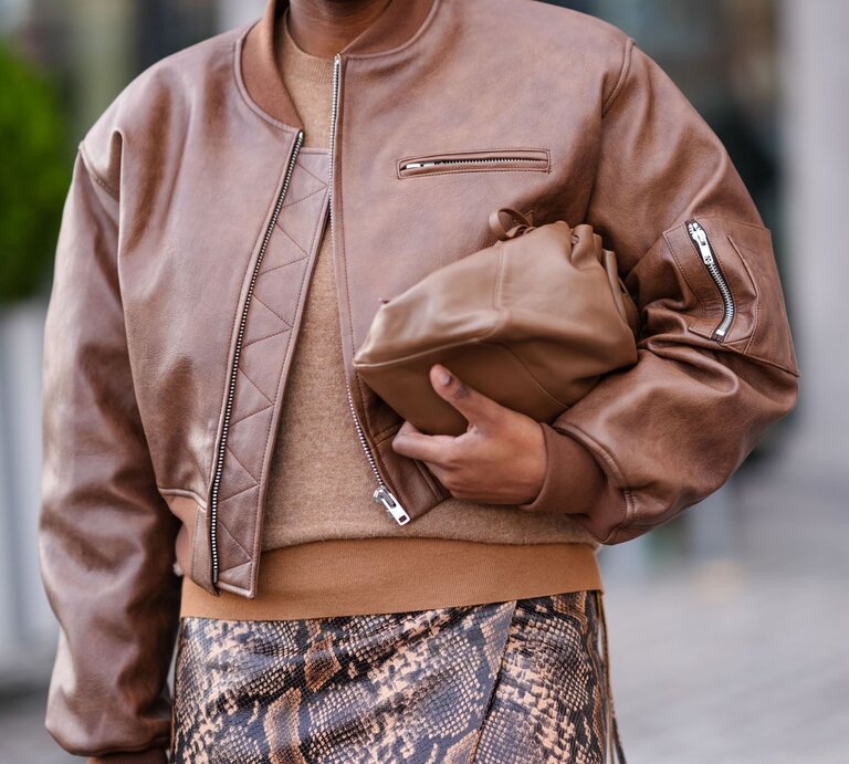 Nahaufnahme eines stylishen Outfits mit brauner Lederjacke, beigem Strick und einem Rock mit Schlangenmuster auf urbanem Hintergrund. | © Edward Berthelot/Getty Images