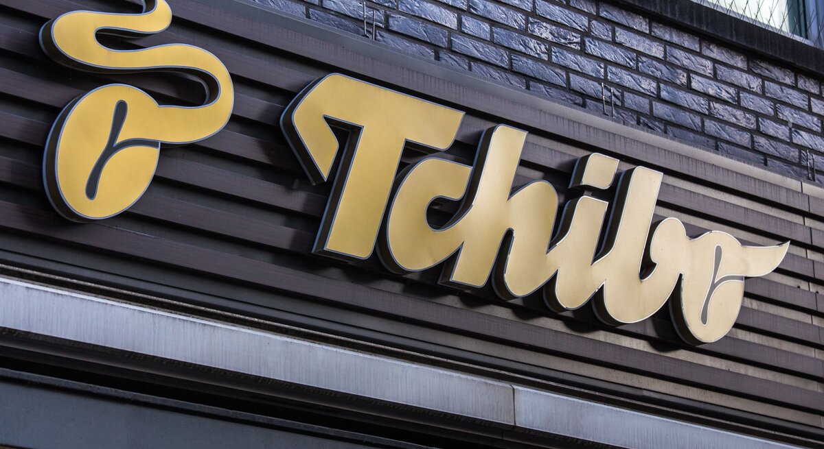Gelbes Tchibo-Logo auf dunkelbrauner Holzverkleidung einer Hausfassade mit grauen Backsteinen im Hintergrund.