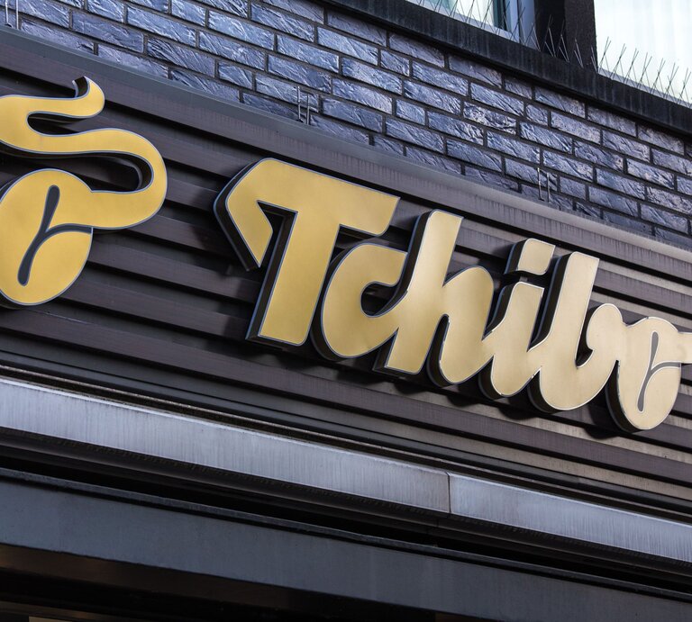 Gelbes Tchibo-Logo auf dunkelbrauner Holzverkleidung einer Hausfassade mit grauen Backsteinen im Hintergrund. | © Adobe Stock/Tobias Arhelger