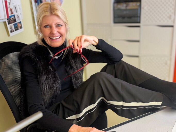 Lachende blonde Renate Zott in schwarzem Outfit mit Fellweste sitzt lässig im Bürostuhl, hält eine rote Brille und blickt freundlich in die Kamera. | © Renate Zott