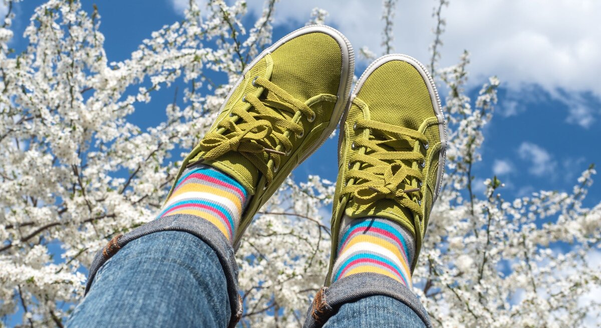 Zwei Beine in hochgeworfenen Jeans mit knallgrünen Sneakers und bunten Ringelsocken vor blühenden Kirschbäumen im Frühlingshimmel.