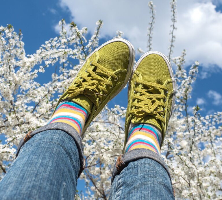 Zwei Beine in hochgeworfenen Jeans mit knallgrünen Sneakers und bunten Ringelsocken vor blühenden Kirschbäumen im Frühlingshimmel. | © Shutterstock/ SvetikovaV