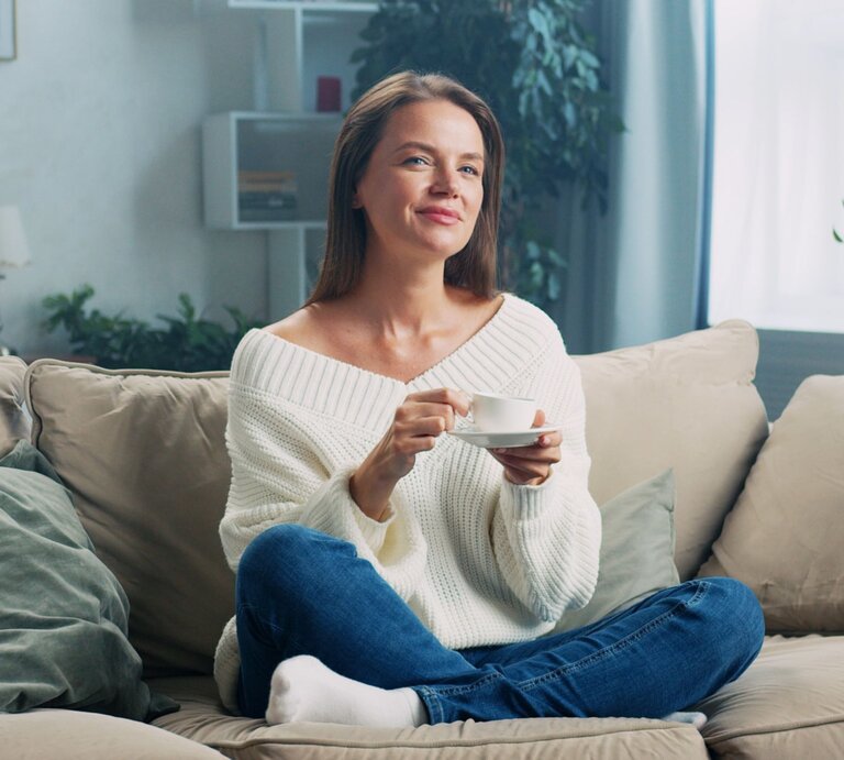 Lächelnde Frau in weißem Strickpullover und Jeans trinkt Kaffee auf einem gemütlichen Sofa in einem hellen Wohnzimmer. | © Adobe Stock/ MIKHAIL