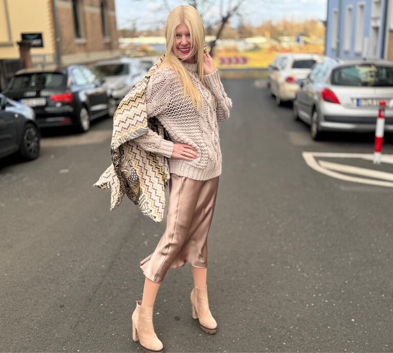 Renate Zott in beigem Strickpullover und glänzendem Satinrock posiert lächelnd in einer ruhigen Straße, umgeben von parkenden Autos. | © Renate Zott