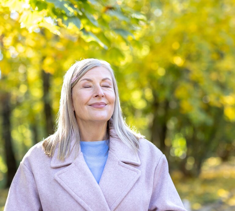 Eine Person mit langen graublonden Haaren trägt einen hellrosa Mantel und einen blauen Pullover im herbstlichen Park. | © Adobe Stock/ Liubomir