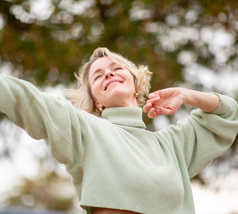 Eine Person mit blondem Haar trägt einen hellgrünen Pullover mit Rollkragen und hebt beide Arme vor einer herbstlichen Baumkulisse. | © Adobe Stock/ Paula