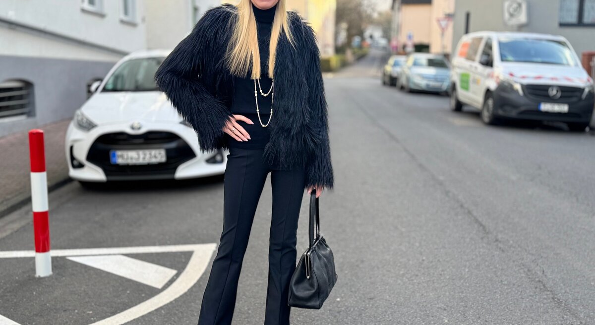 Eine blonde Frau in schwarzer Kleidung mit Pelzjacke, Mütze und Handtasche lächelt auf einer Straße mit geparkten Autos.
