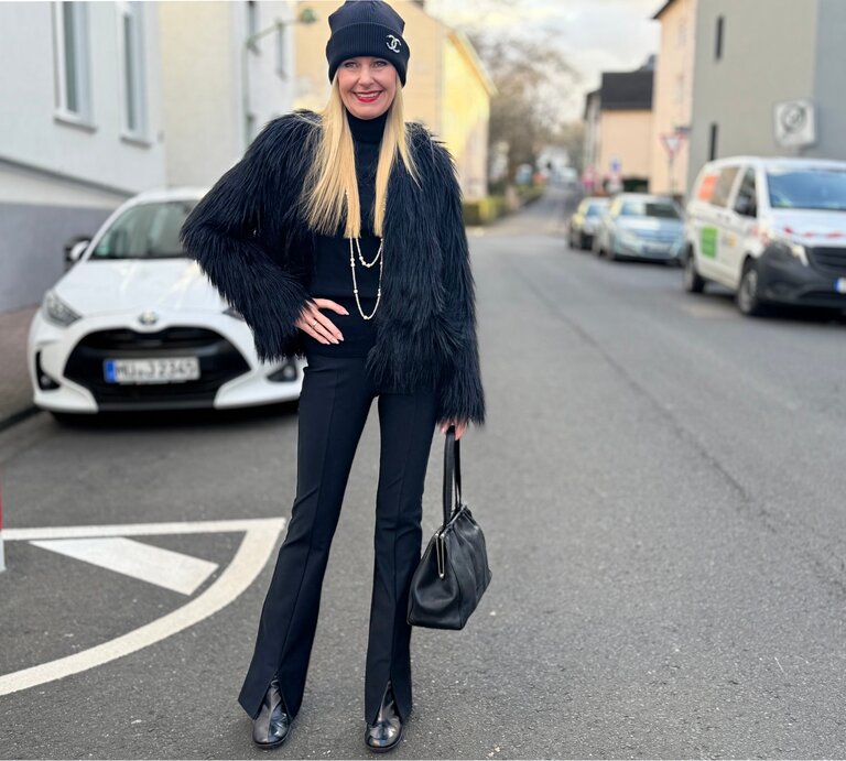 Eine blonde Frau in schwarzer Kleidung mit Pelzjacke, Mütze und Handtasche lächelt auf einer Straße mit geparkten Autos. | © Renate Zott