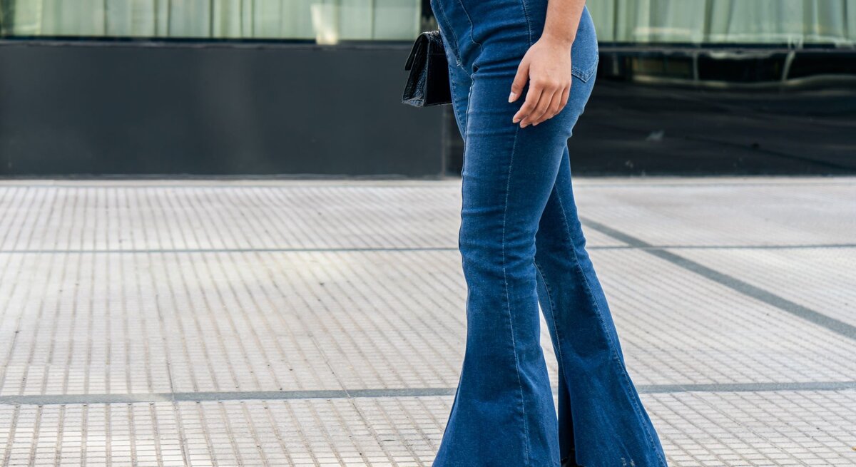 Eine Frau in einem karierten Hemd und blauen Schlaghosen läuft auf einem modernen Gehweg und trägt eine schwarze Umhängetasche.