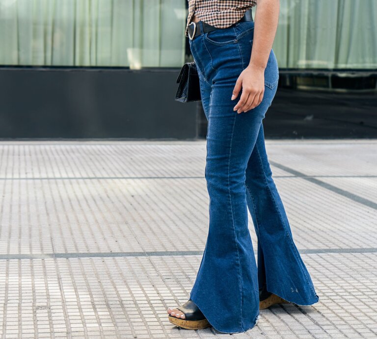 Eine Frau in einem karierten Hemd und blauen Schlaghosen läuft auf einem modernen Gehweg und trägt eine schwarze Umhängetasche. | © Adobe Stock/ HC FOTOSTUDIO