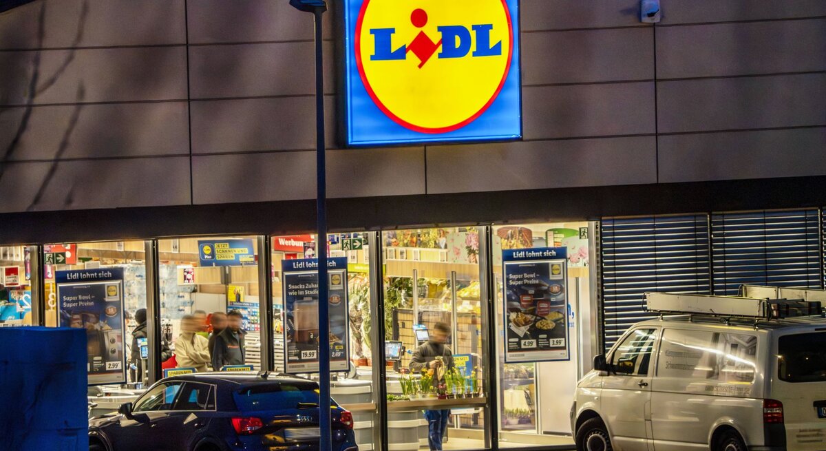  Auf dem Foto ist der beleuchtete Eingangsbereich einer Lidl-Filiale während der Abend- oder Nachtzeit zu sehen. Das markante Lidl-Logo mit gelbem Hintergrund und rotem Schriftzug leuchtet hell über dem Eingang und zieht die Blicke auf sich. Vor dem Supermarkt parken Autos, und man kann Personen erkennen, die sich im Inneren des Geschäfts bewegen. Die Umgebung ist durch die Beleuchtung des Ladens und die Dämmerung oder die nächtliche Umgebung dunkel kontrastiert.
