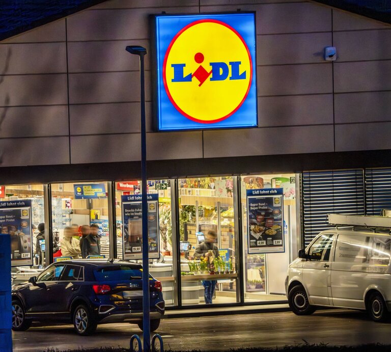  Auf dem Foto ist der beleuchtete Eingangsbereich einer Lidl-Filiale während der Abend- oder Nachtzeit zu sehen. Das markante Lidl-Logo mit gelbem Hintergrund und rotem Schriftzug leuchtet hell über dem Eingang und zieht die Blicke auf sich. Vor dem Supermarkt parken Autos, und man kann Personen erkennen, die sich im Inneren des Geschäfts bewegen. Die Umgebung ist durch die Beleuchtung des Ladens und die Dämmerung oder die nächtliche Umgebung dunkel kontrastiert. | © IMAGO / Wolfgang Maria Weber