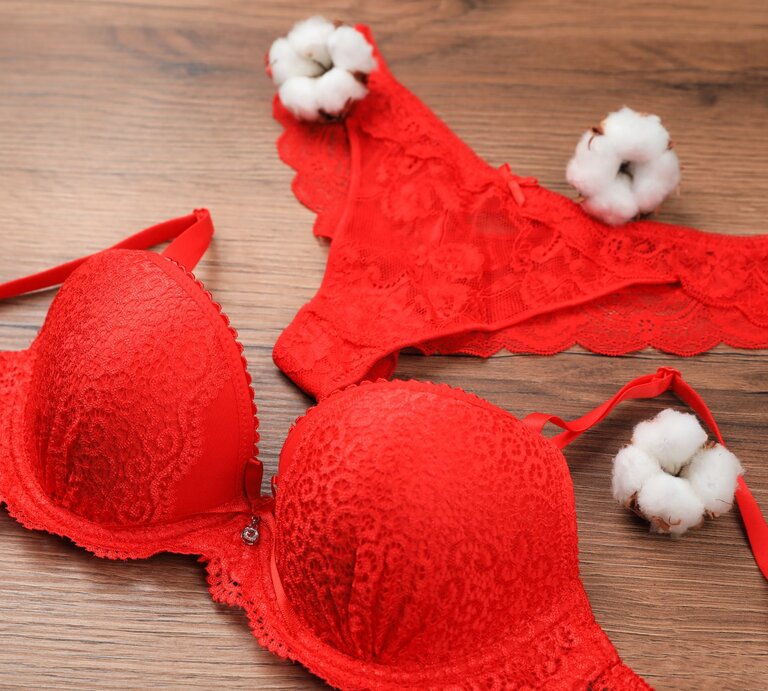 Rote Spitzenunterwäsche bestehend aus einem BH und einem Slip liegt auf einem Holztisch, dekoriert mit weißen Baumwollblüten. | © Getty Images / Liudmila Chernetska