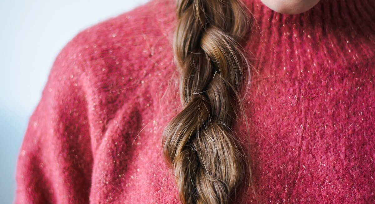 Eine Frau mit geflochtenem braunem Haar trägt einen rosafarbenen Glitzerpullover und dezenten Lippenstift.