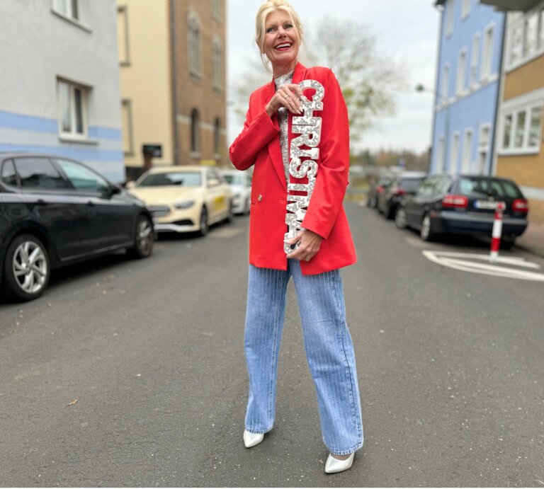 Eine blonde Frau in rotem Blazer und hellblauen Jeans lächelt auf einer Straße und hält eine silberne „Christmas“-Dekoration. | © Renate Zott