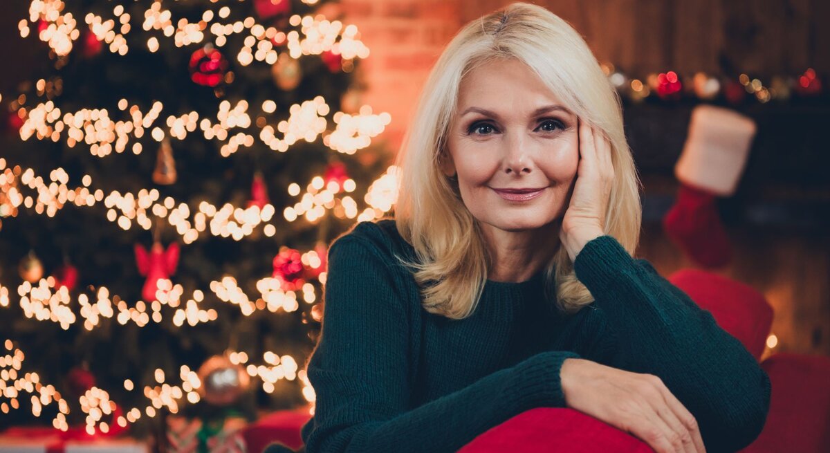 Eine lächelnde blonde Frau mittleren Alters lehnt in einem grünen Pullover entspannt auf einem roten Sofa, vor einem leuchtenden Weihnachtsbaum.