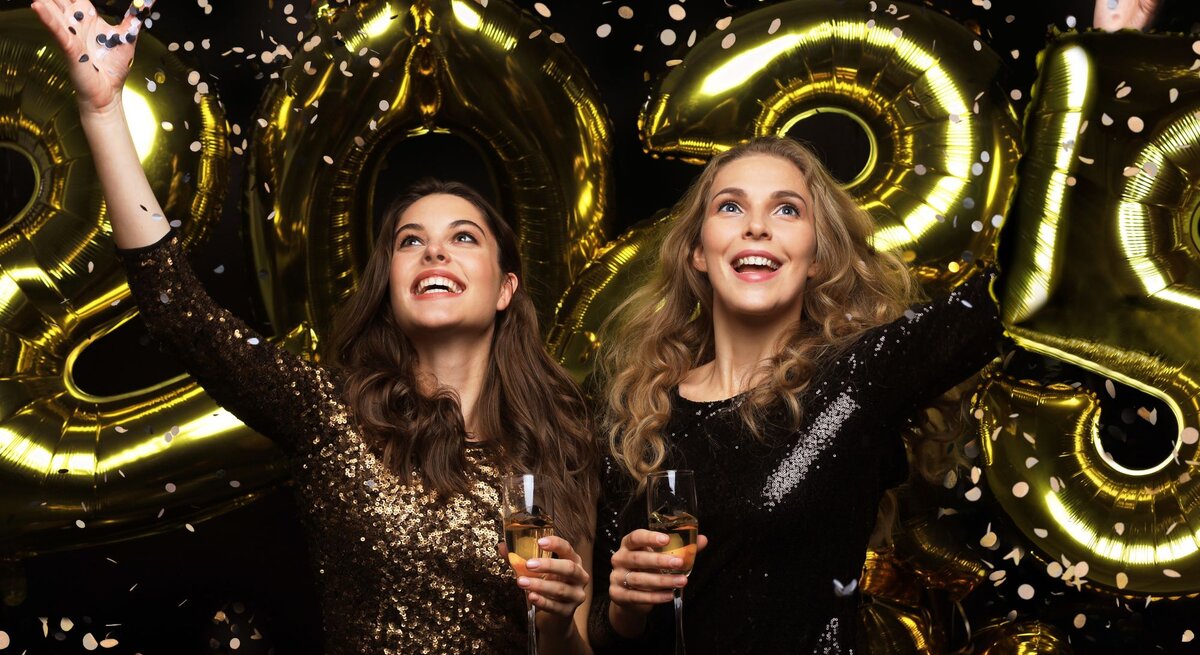 Zwei junge Frauen in glitzernden Pailletten-Kleidern von Lidl feiern fröhlich mit Sektgläsern und goldenen Ballons vor schwarzem Hintergrund.