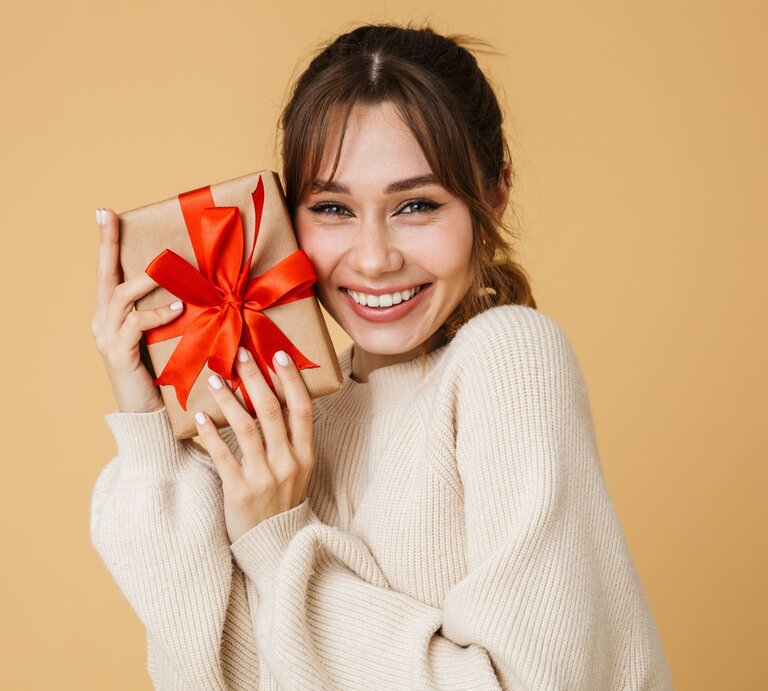 Junge Frau mit braunem Haar und beigefarbenem Pullover hält lächelnd ein Geschenk mit roter Schleife vor beigem Hintergrund. | © Adobe Stock/ Drobot Dean