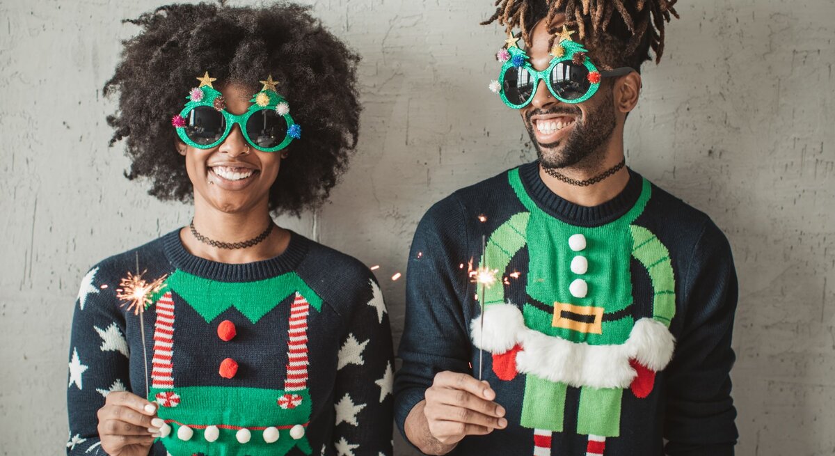 Zwei fröhliche Menschen in bunten Weihnachtspullovern und auffälligen Brillen mit Sternen und Weihnachtsdekor, halten Wunderkerzen vor grauer Wand.
