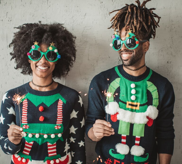 Zwei fröhliche Menschen in bunten Weihnachtspullovern und auffälligen Brillen mit Sternen und Weihnachtsdekor, halten Wunderkerzen vor grauer Wand. | © Getty Images/ svetikd