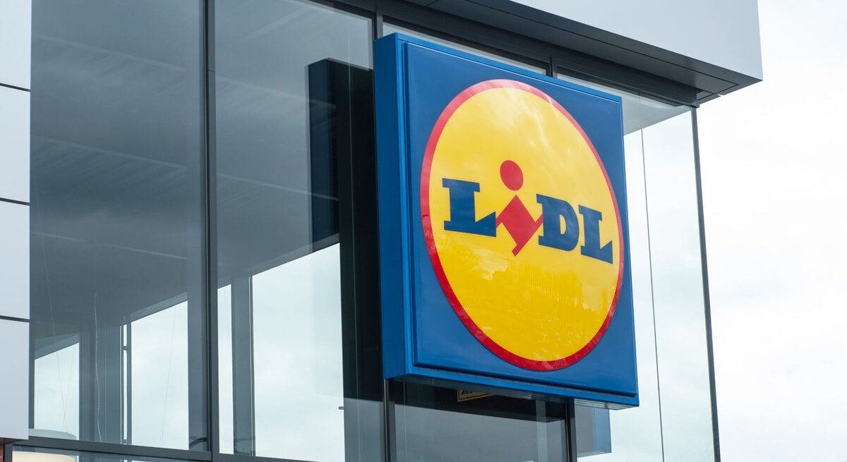  Das Bild zeigt das typische Logo des Einzelhändlers Lidl an einem Geschäftsgebäude. Die moderne Architektur des Gebäudes mit großen Glasflächen und der klare Himmel im Hintergrund vermitteln einen zeitgenössischen Eindruck. Lidl ist bekannt für sein Discount-Supermarktkonzept, das in vielen Ländern vertreten ist und eine breite Palette von Produkten, darunter Lebensmittel, Haushaltswaren und saisonale Artikel, zu günstigen Preisen anbietet. Das präsentierte Logo mit dem blauen Hintergrund und dem gelben Kreis mit roter Schrift ist sofort erkennbar und repräsentiert die Marke Lidl weltweit.
